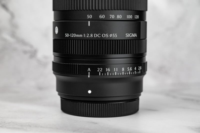 A close-up view of a black camera lens barrel showing focal length markings from 50 to 120mm, aperture settings from 2.8 to 22, and the text "SIGMA 50-120mm 1:2.8 DC OS".