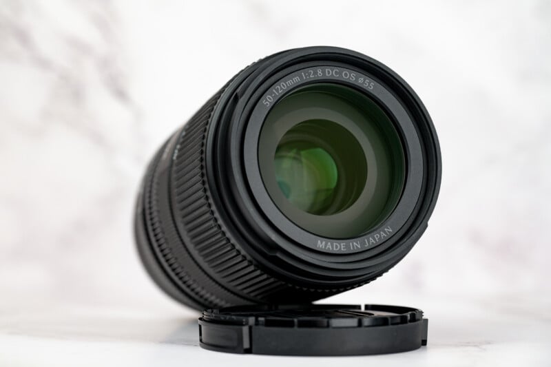 A black camera lens with a 50-120mm focal range and 1:2.8 aperture sits upright on a white marble surface next to its detached lens cap.