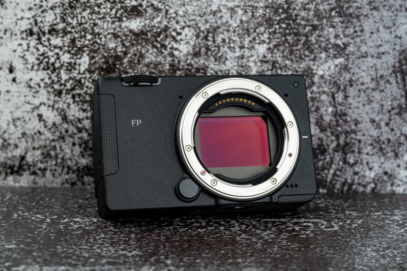 A black digital camera body with a prominent lens mount and visible sensor sits on a textured, mottled gray surface.