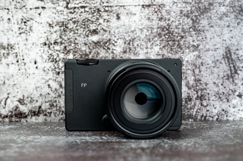 A black digital camera with a lens attached sits on a textured surface against a mottled grey background, with the letters "FP" printed on the front.