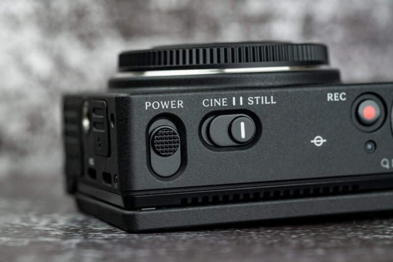A close-up view of the top panel of a black camera, showing a power switch, a mode selector switch labeled CINE and STILL, and a red record button.