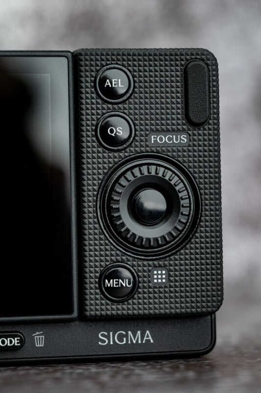 Close-up of the back of a black Sigma camera, showing buttons labeled AEL, QS, FOCUS, and MENU, along with a central control dial and a trash icon.