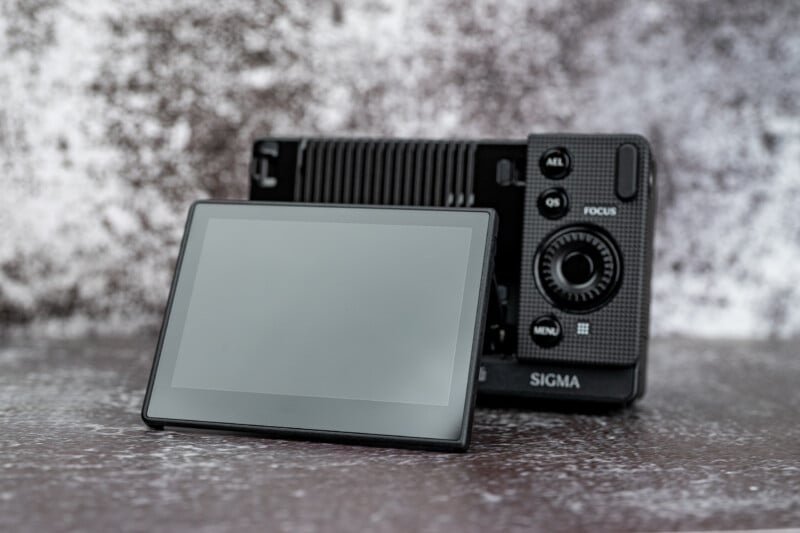 A black digital camera with a detached, tilted screen sits on a textured surface against a mottled gray background.