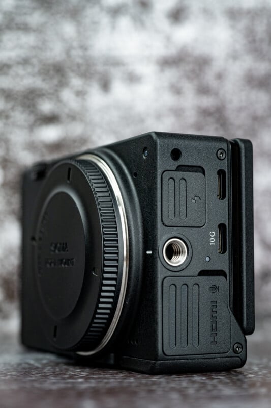 A side view of a black digital camera body with a lens mount cap, a tripod mounting hole, and several ports labeled with icons and text including 10G and HDMI.