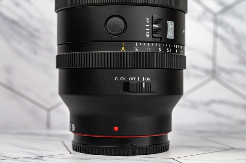 A close-up of a camera lens barrel showing an aperture ring, an AF/MF switch, and a click on/off switch against a marble background.
