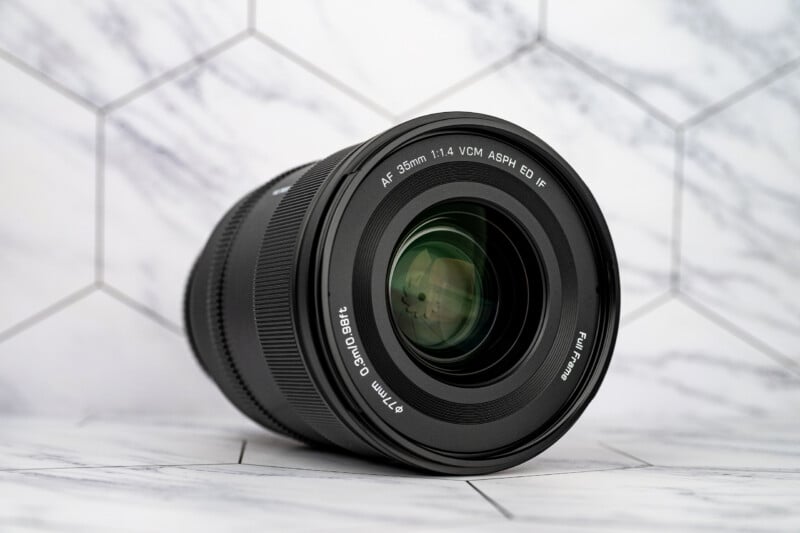 A black camera lens with a 35mm focal length and 1.4 aperture sits on a white marble surface with a hexagonal tile pattern background.