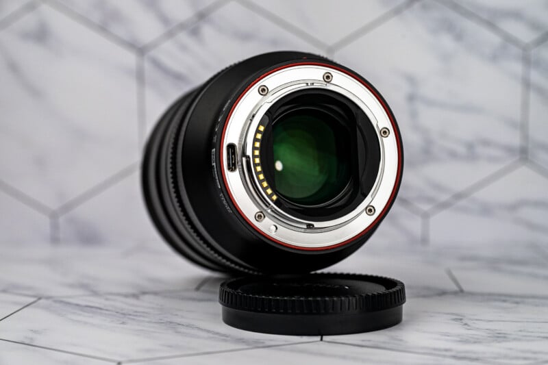 A camera lens with a metal mount and electronic contacts sits on a marble-patterned surface next to its black lens cap.