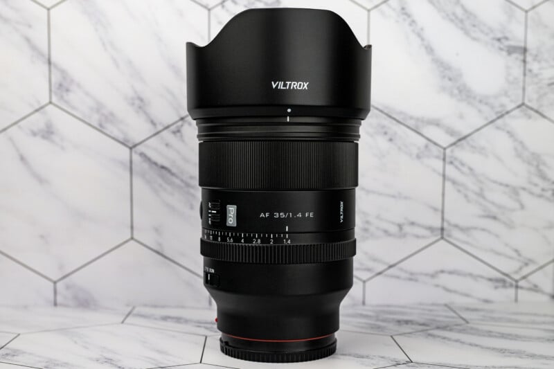 A Viltrox AF 35/1.4 FE camera lens stands upright against a white marble-patterned hexagonal tile background.