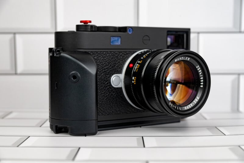 A black Leica M-series rangefinder camera with a large hand grip and a lens labeled "Mandler" sits on a white subway tile surface.