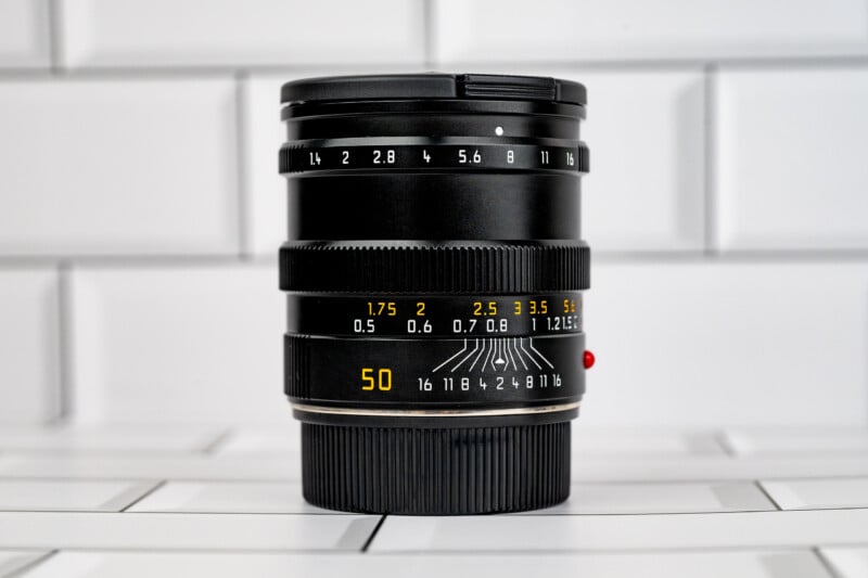 A black 50mm camera lens stands upright on a white tiled surface, featuring white aperture markings and yellow distance scales.