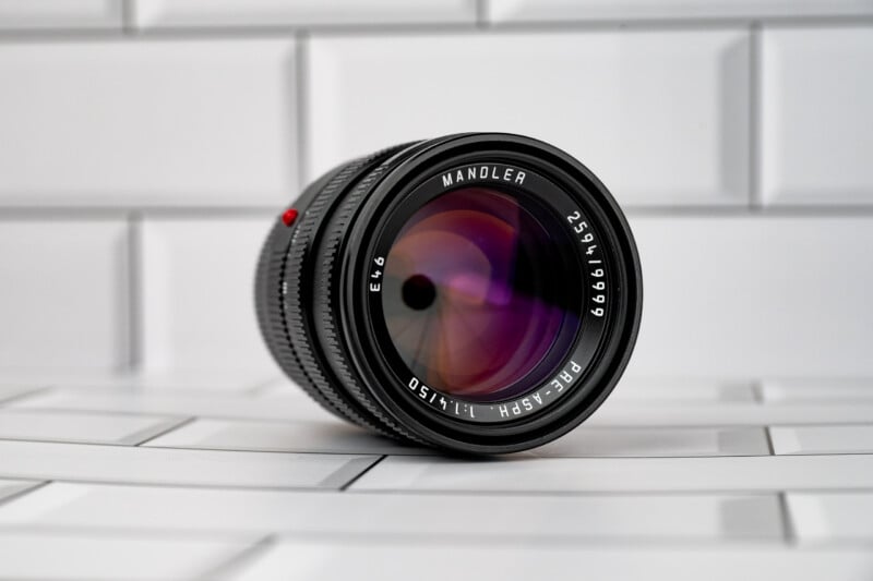 A black camera lens with the text "MANDLER", "E46", "2594/19999", and "PRE-ASPH. 1:1.4/50" printed on the front, resting on a white tiled surface.