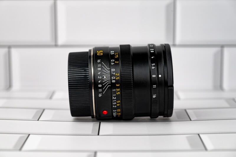 A black manual focus camera lens with yellow distance and aperture markings sits horizontally on a white tiled surface.