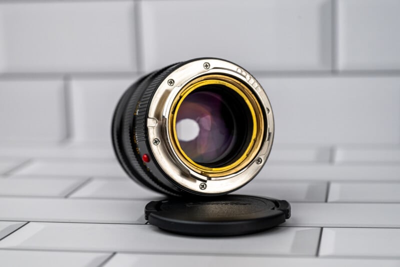 A camera lens with a silver mount and gold-colored inner ring sits on a white tiled surface next to its black lens cap.