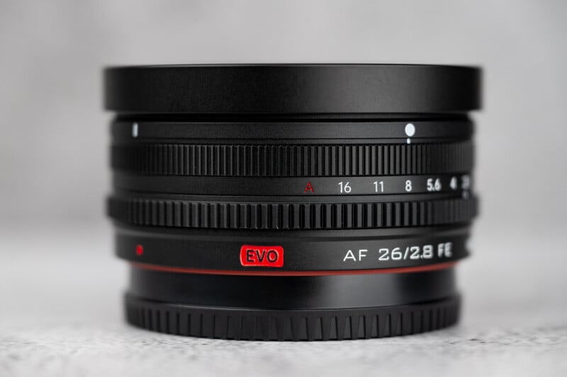 A black camera lens stands upright on a textured grey surface, featuring an aperture ring with markings from 16 to 5.6, an "EVO" logo in a red box, and the text "AF 26/2.8 FE" printed on the side.