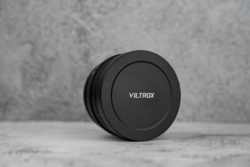 A black Viltrox camera lens cap is attached to a lens, resting on a textured gray surface against a matching gray background.