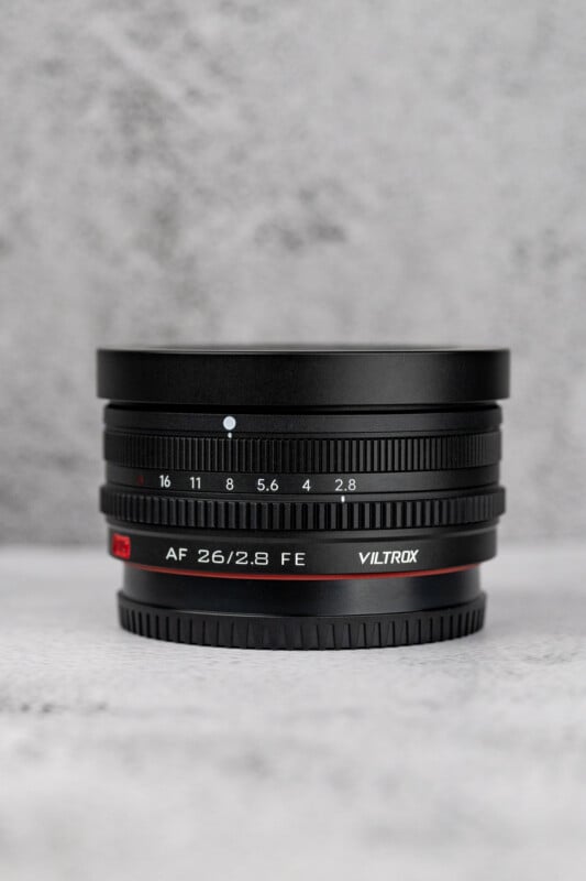 A black Viltrox AF 26/2.8 FE camera lens sits upright on a light gray textured surface against a matching gray background.