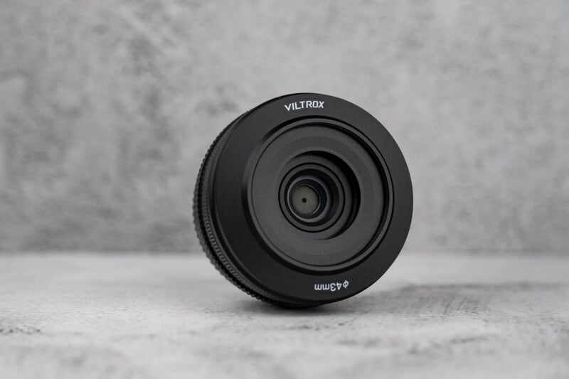 A black Viltrox camera lens with a 43mm filter thread size sits upright on a textured gray surface.