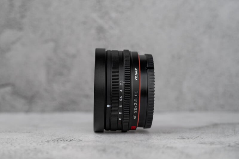 A black Viltrox AF 26/2.8 FE camera lens stands upright on a textured gray surface against a matching gray background.