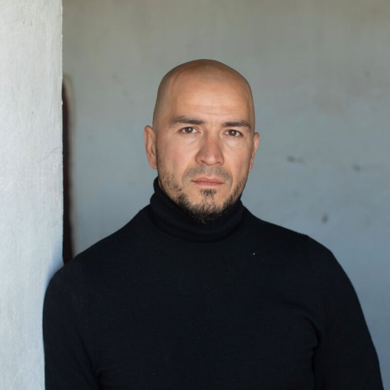 A bald man with a short beard and a black turtleneck stands against a plain wall.