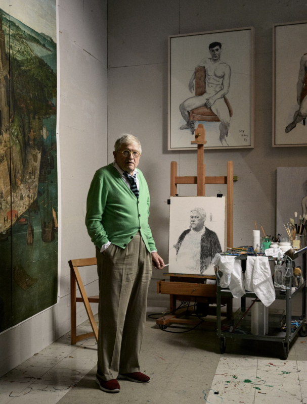 An elderly man with glasses and a green cardigan stands in an art studio next to an easel holding a charcoal self-portrait.
