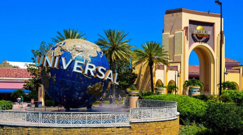 The iconic Universal Studios globe sculpture sits in a fountain in front of a large, tan-colored archway under a clear blue sky.