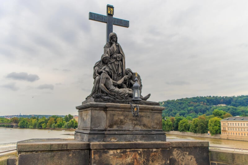 A bronze sculpture of the Pietà featuring Jesus and mourning figures stands on a stone pedestal atop a bridge, with a river and trees in the background.