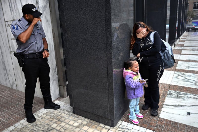 A security guard and a woman with a young child stand against a large pillar, all three appearing to be crying.
