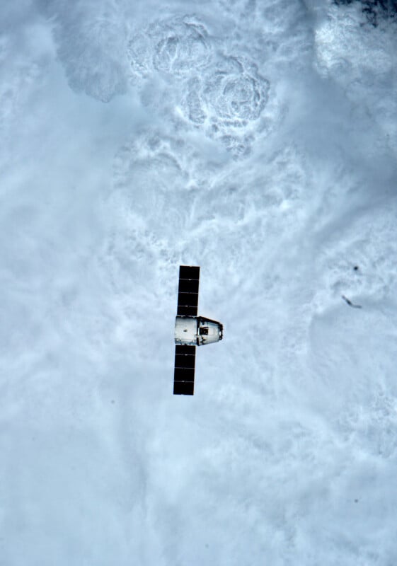 A SpaceX Dragon spacecraft with extended solar arrays floats in space above a dense layer of white clouds.