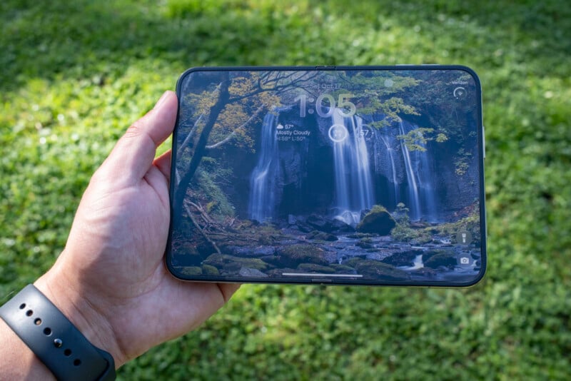 A hand holds a foldable smartphone displaying a lock screen with a waterfall wallpaper, the time 1:05, and weather widgets.