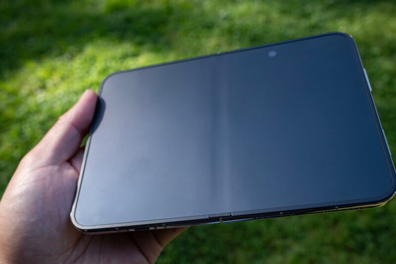 A hand holds an unfolded, black foldable smartphone against a blurred green grass background.