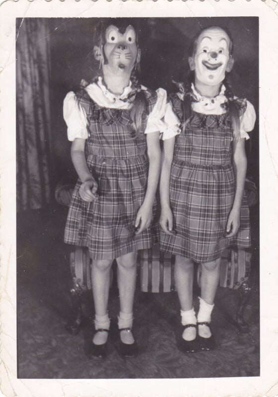 Two young girls wearing matching plaid dresses and masks stand side-by-side in front of a chair.
