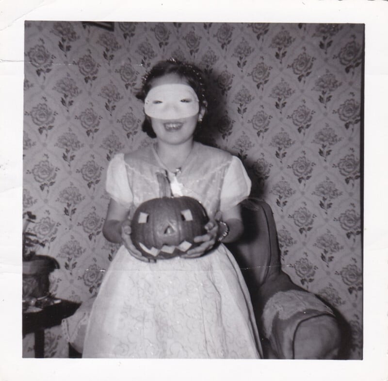 A young girl in a light-colored dress and a plain white mask holds a small carved pumpkin while sitting in a chair against floral wallpaper.
