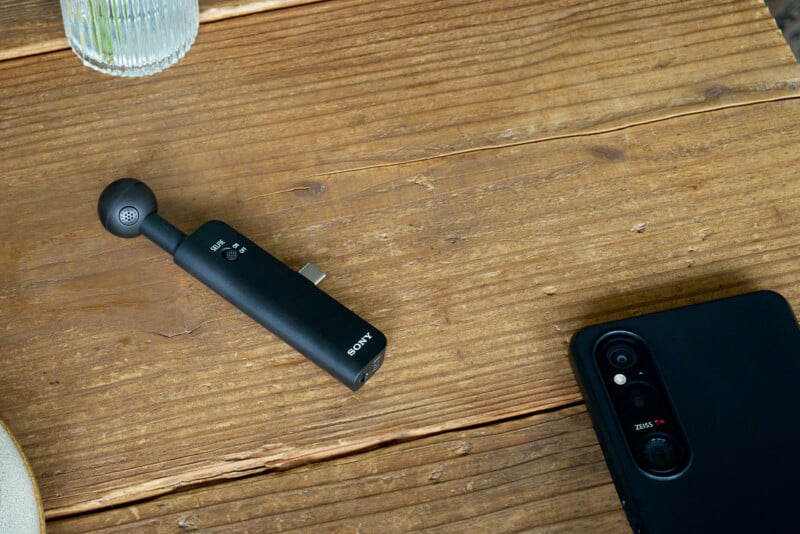 A black Sony-branded microphone attachment with a selfie switch sits on a wooden table next to a smartphone with a Zeiss camera lens.