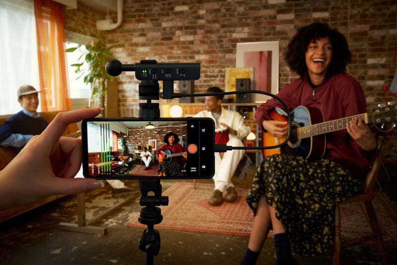 A hand holds a smartphone on a tripod, recording a woman playing acoustic guitar while two other people sit in the background.