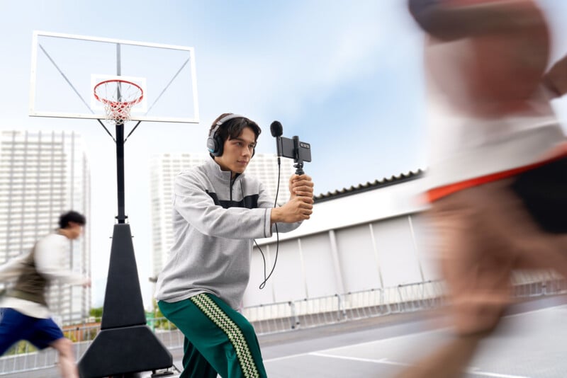 A young man wearing headphones holds a smartphone on a gimbal with a microphone attached while standing on an outdoor basketball court, with blurred figures of people running past him.