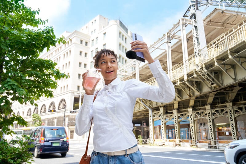 A young woman with curly hair, wearing a white button-down shirt and jeans, holds a pink drink and a smartphone with a microphone attachment while standing on a city street near an elevated train track.