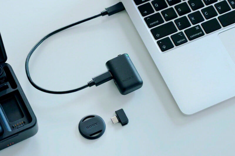 A Sony wireless microphone receiver is connected to a laptop via a USB-C cable, with a circular magnetic mount and a USB-C adapter lying on the desk nearby.