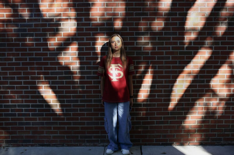 A young woman with long hair stands against a brick wall, wearing a maroon Florida State University t-shirt and blue cargo pants.