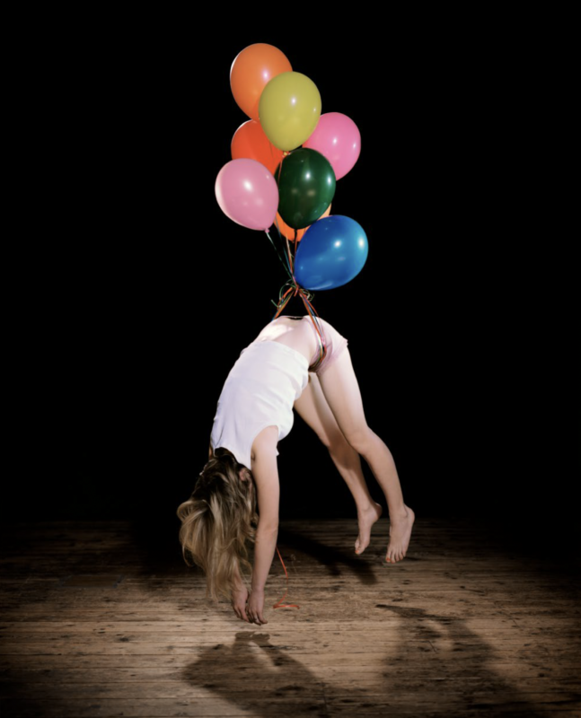 A person in a white tank top and shorts is suspended upside down, held by a bunch of colorful balloons tied around their waist.