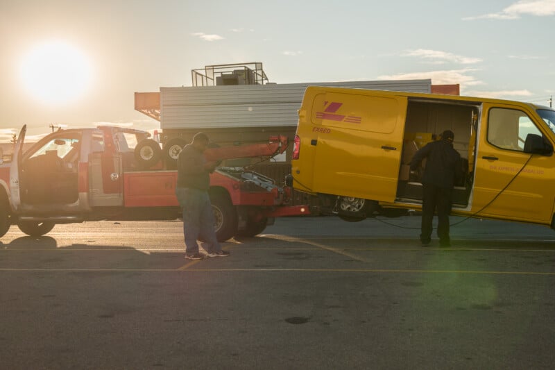 A tow truck is hooked up to a yellow DHL Express delivery van in a parking lot at sunset, with two men standing nearby.