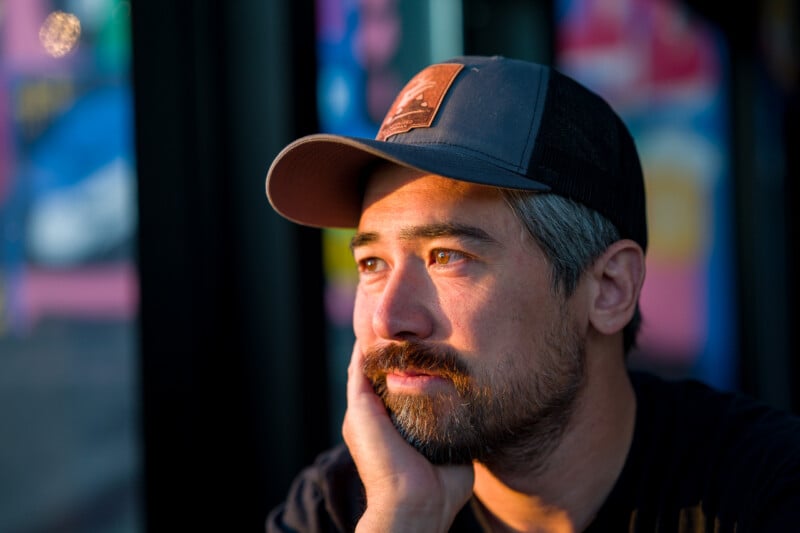 A man with a beard and graying hair wears a baseball cap while resting his chin on his hand, illuminated by warm sunlight.