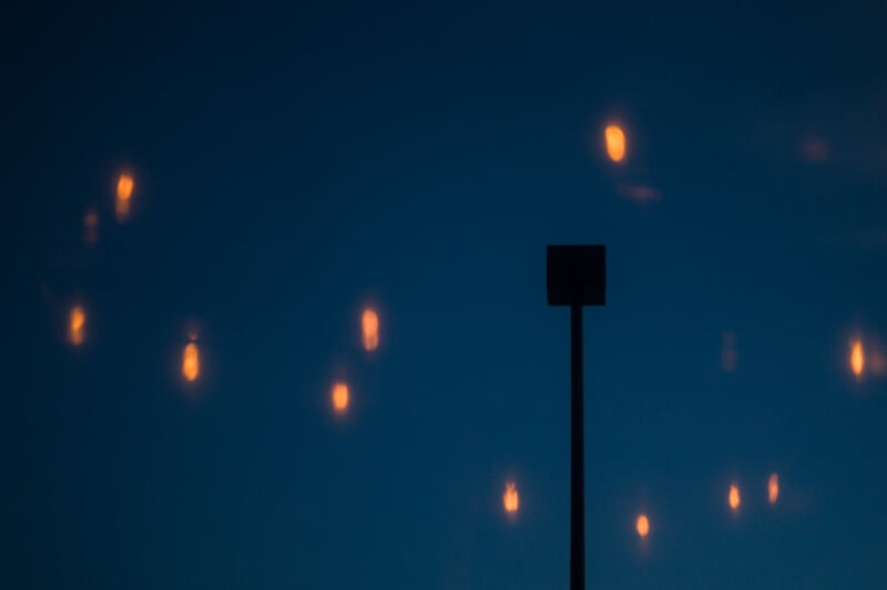 A dark silhouette of a square-topped pole stands against a deep blue sky filled with scattered, out-of-focus orange lights.