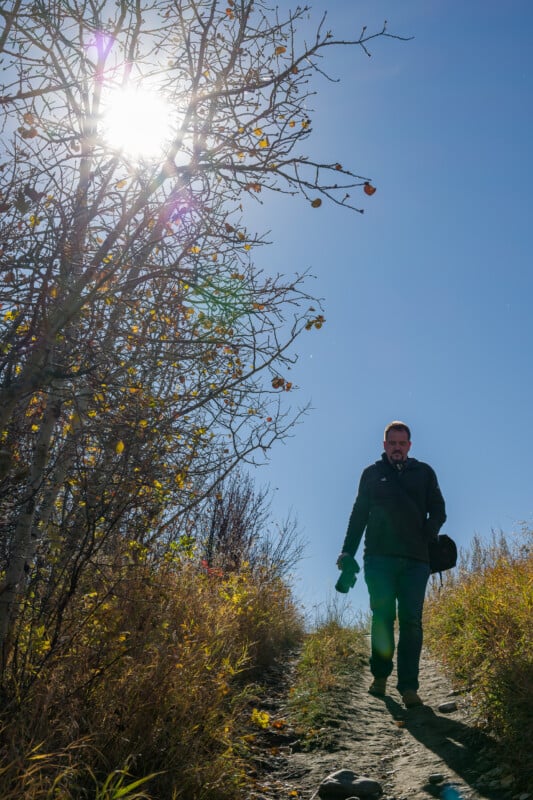 A man carrying a camera walks down a dirt path through tall, golden grasses under a bright sun and clear blue sky.