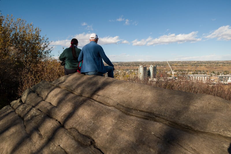 A man and a woman sit on a large rock overlooking a city landscape under a blue sky.