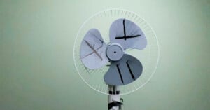 A white pedestal fan with three grey blades, each marked with a unique black hand-drawn symbol, mounted against a plain wall.