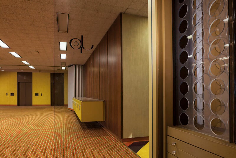 An interior view of a retro office lobby featuring patterned orange carpet, yellow walls, wood-paneled partitions, and a decorative screen with circular cutouts.