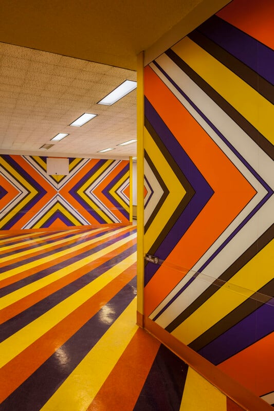 A corner of a room features walls and floors covered in a repeating, vibrant chevron pattern of orange, yellow, purple, and white stripes.