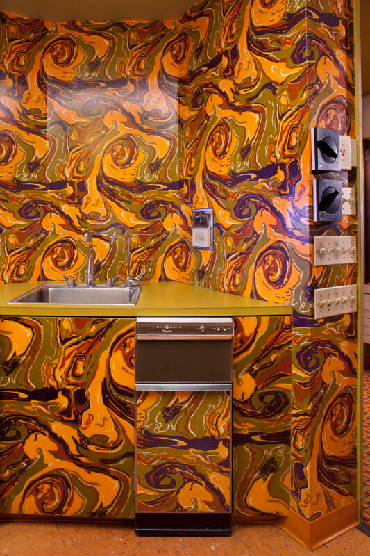 A kitchen corner features a sink and a General Electric trash compactor, both covered in a swirling, psychedelic orange and brown pattern that matches the surrounding walls.