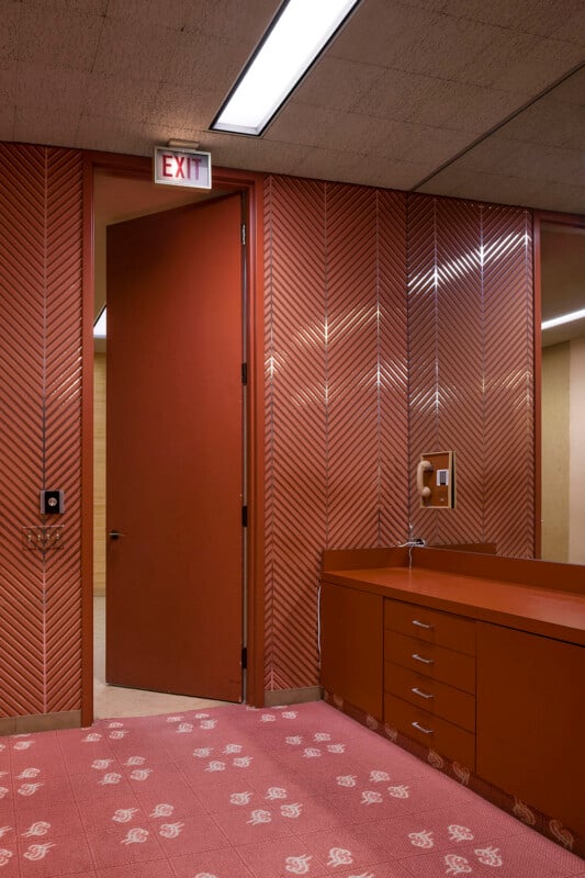 An interior view of a room with terracotta-colored herringbone walls, a matching door with an exit sign above, and a patterned carpet.