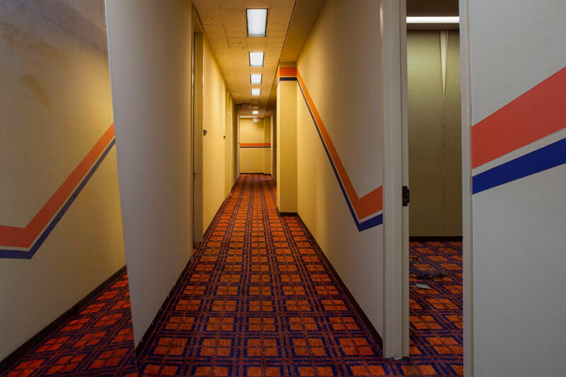 A long, narrow hotel hallway with patterned orange and blue carpet, yellow walls featuring a matching orange and blue stripe, and recessed ceiling lights.